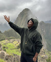 Person in dark green hoodie raising an arm and smiling at Machu Picchu with terraces and Huayna Picchu peak behind under cloudy sky