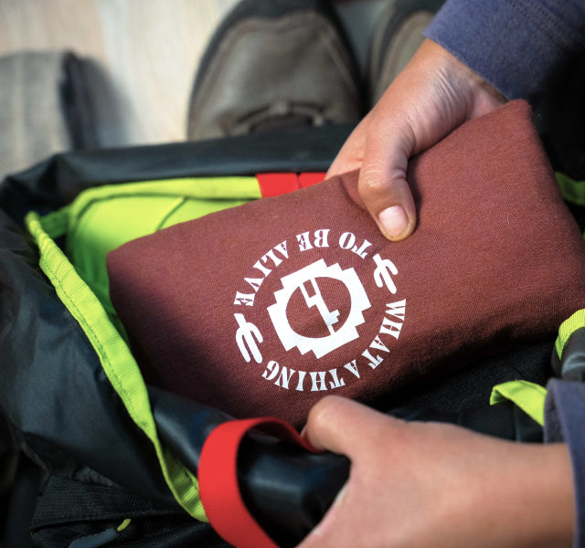 Hands placing a folded clay alpaca base layer with white circular logo into an open hiking backpack