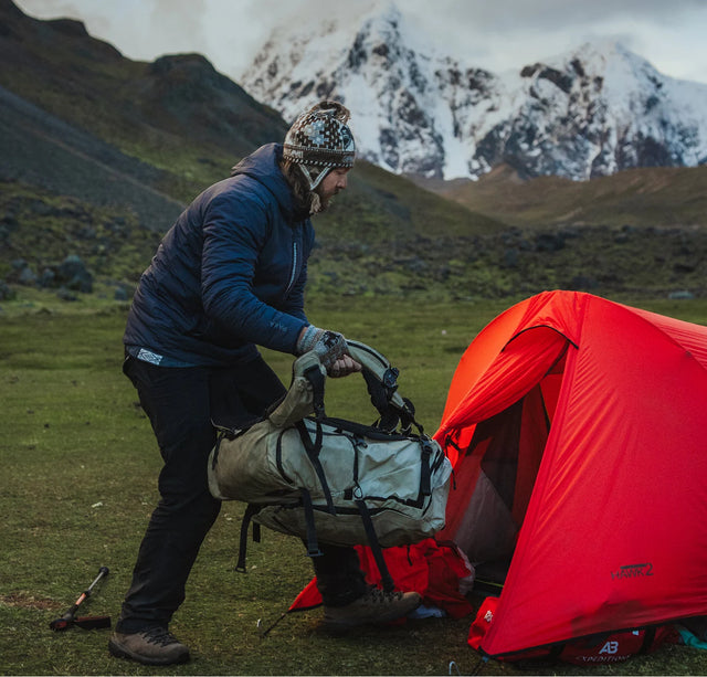 Hiker packing gear beside a red tent with snow-covered mountains in the background