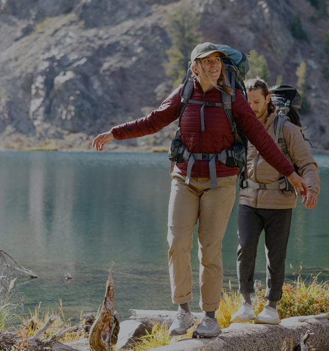 Model wearing Mayu ultralight jacket standing on a rocky lakeshore with lake and mountains in the background