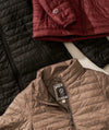 Angled view of Mayu ultralight quilted jacket showing PAKAFILL baffles and zipper with maroon and black jackets behind
