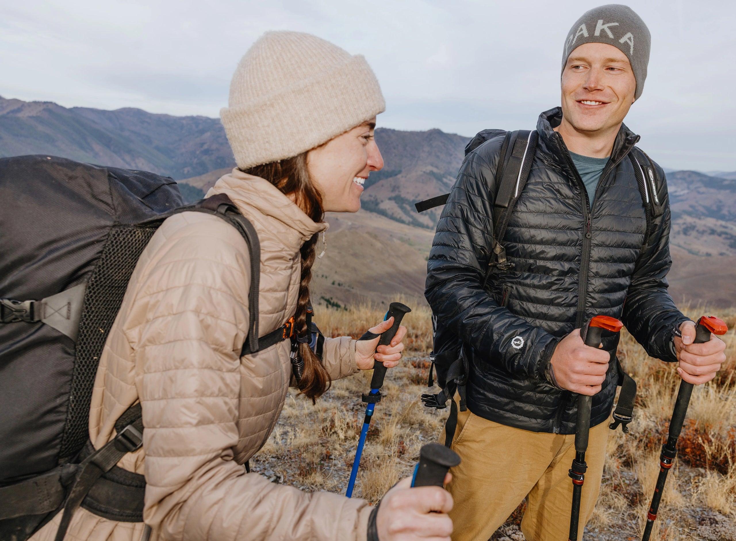 Two hikers on a mountain trail, woman in a beige slim-fit insulated jacket and man in a black ultralight insulated jacket