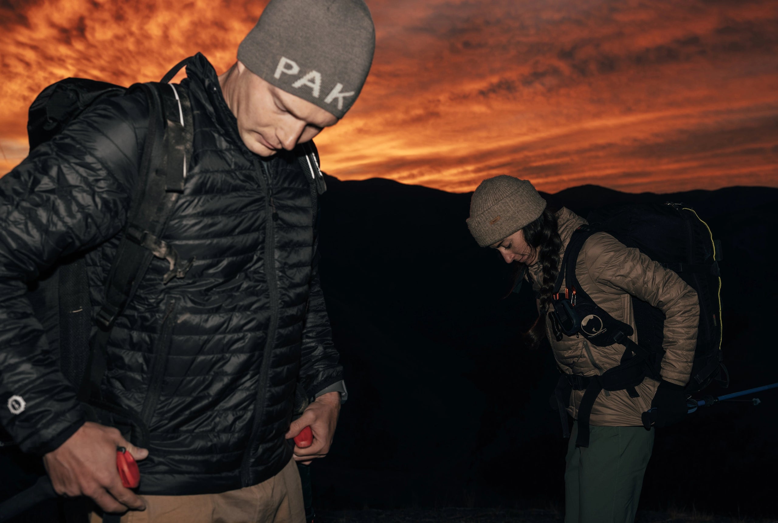 Two people wearing dark Mayu Ultralight jackets standing on a rocky coastline at sunset