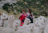 Two people sitting on a rocky hillside, one wearing a red jacket, in a mountainous outdoor landscape