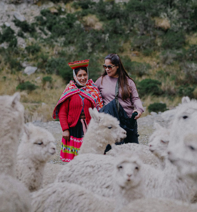Gisella sitting on a rocky hillside with a Peruvian weaver in traditional red clothing, mountains behind them
