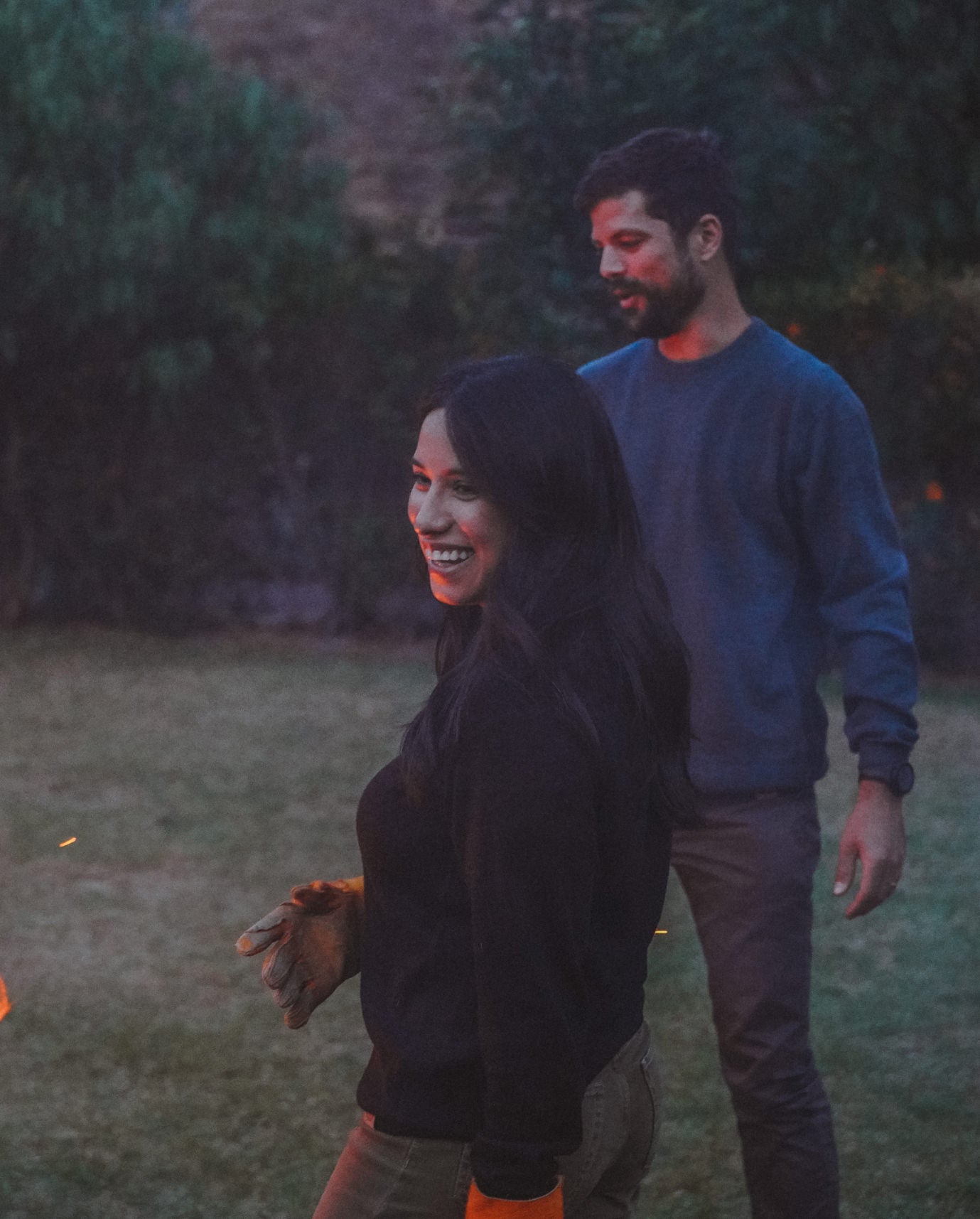 Woman wearing a black Lightweight Crew alpaca crewneck sweater standing beside a small campfire in a backyard, man wearing a blue sweater behind her
