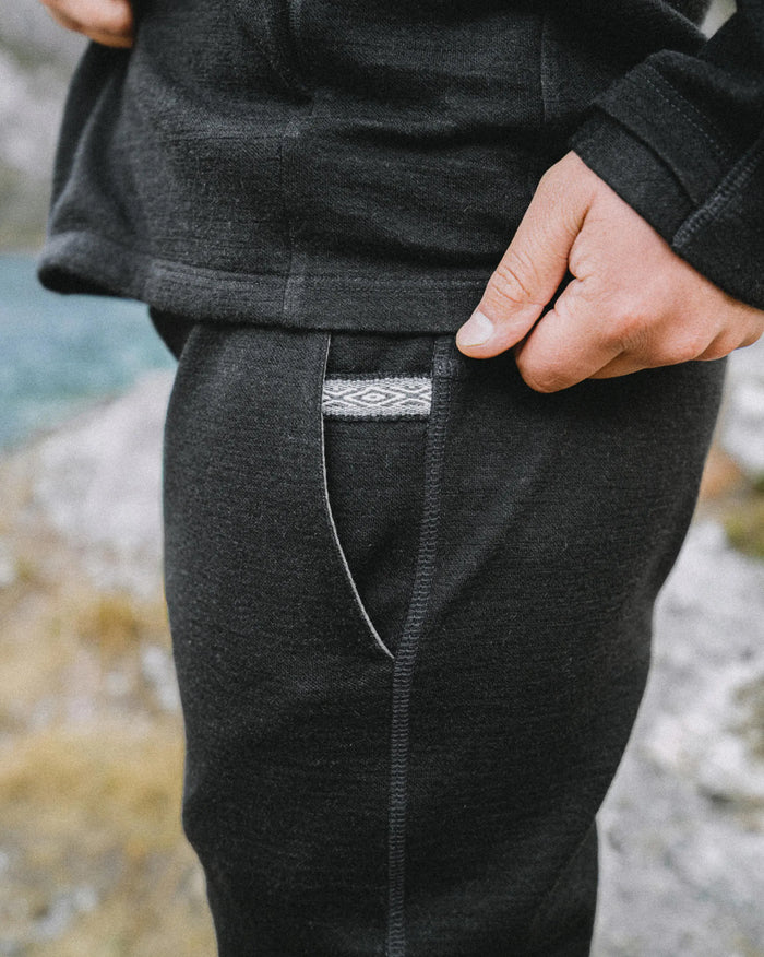 Close-up side view of Men’s Breathe Joggers showing side pocket and woven logo tape on black alpaca terry, blurred rocky background