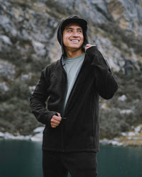Man zipping black Men’s Breathe full-zip hoodie with hood up, slim athletic fit, standing by a mountain lake