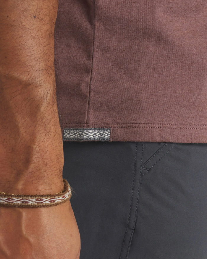 Side hem of men’s Essential Tee in mauve showing woven Inca ID tag and model’s wrist against dark pants