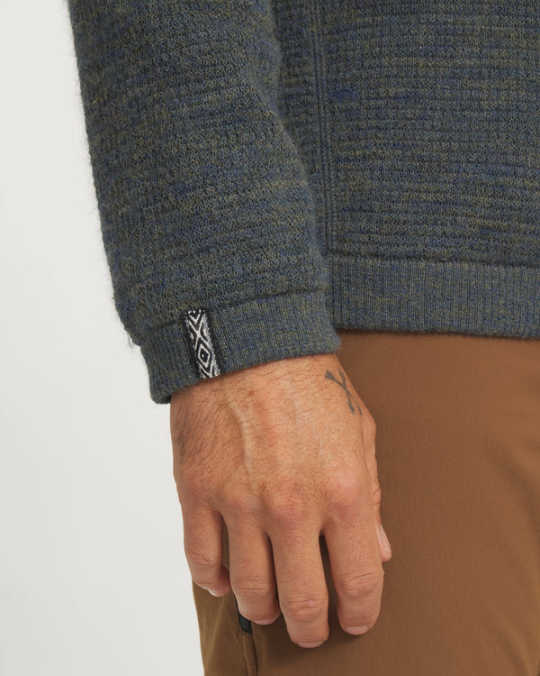 Close-up of a person wearing a textured green sweater with a brand logo on the sleeve against a white background.