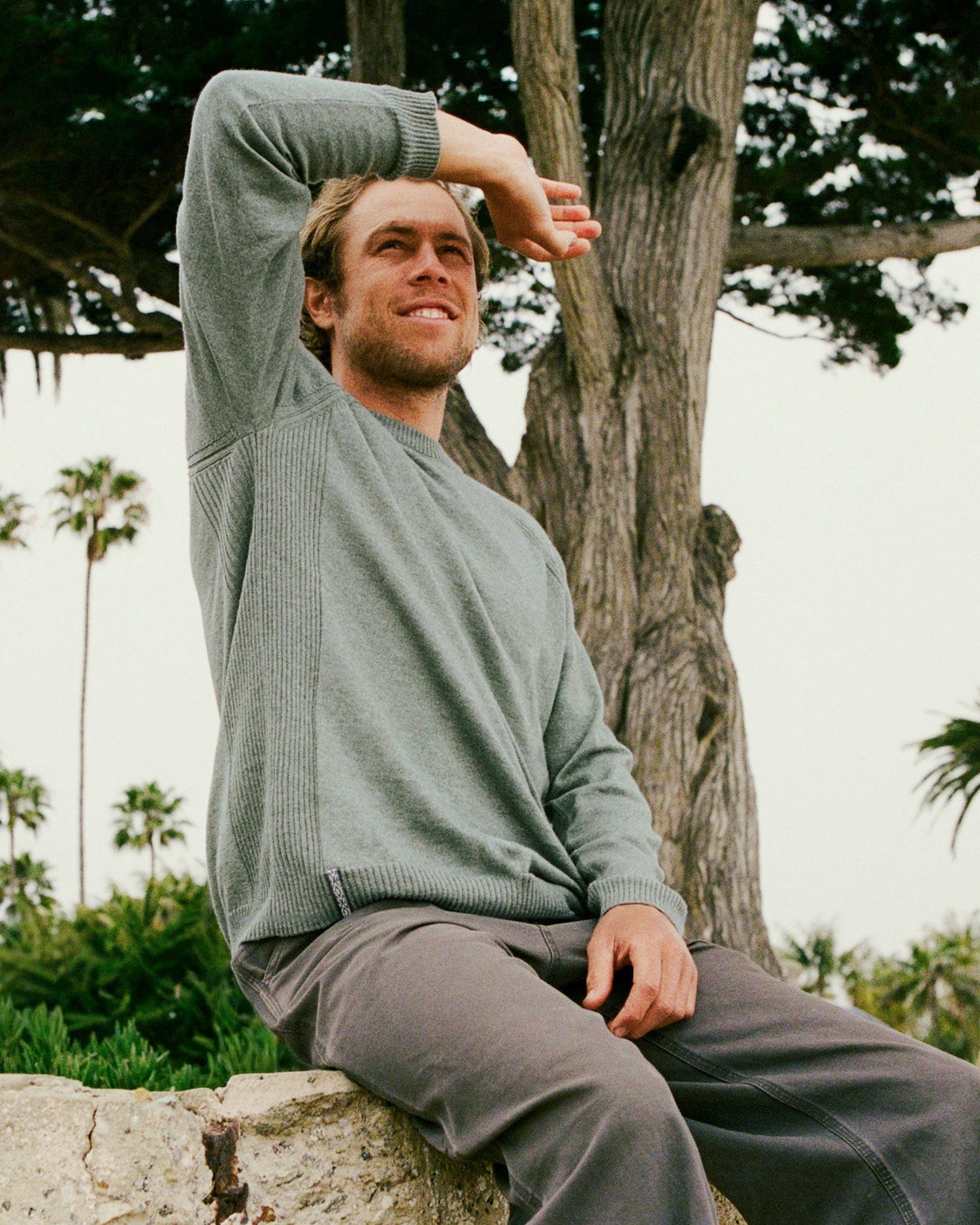 Man sitting outdoors with arms raised, wearing a green sweater and gray pants.