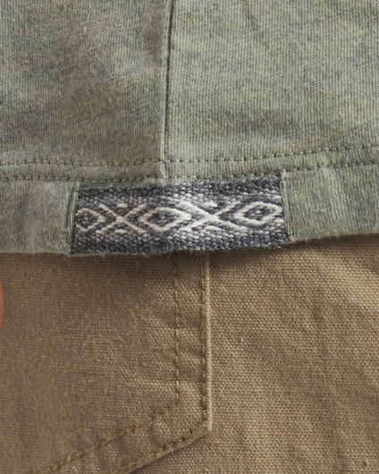 Close-up of Men’s Essential Tee showcasing woven Inca ID hem tag and knit fabric texture with tan pants pocket background