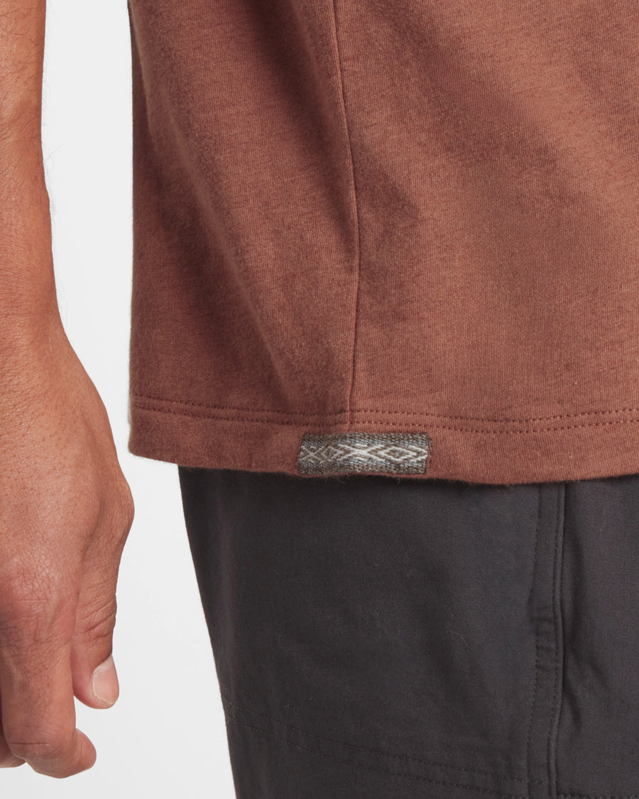 Close up of Men’s Essential Tee hem in cinnamon showing side seam and woven logo tab with model’s hand and black shorts