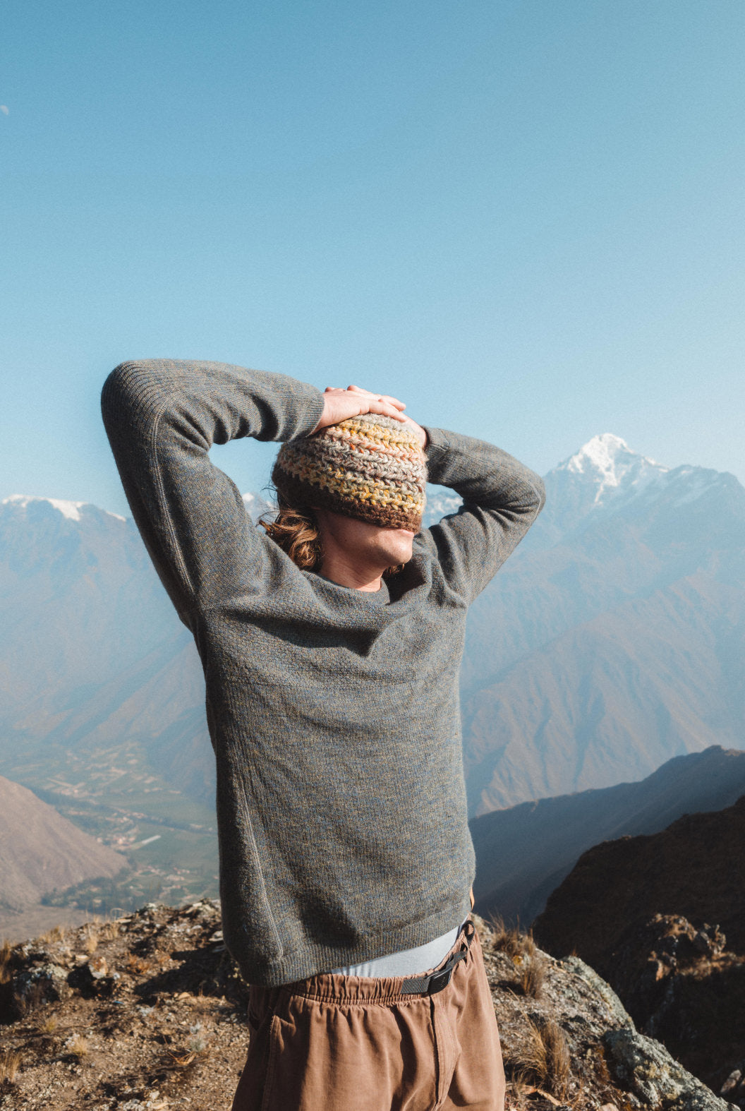 Man on a mountain ridge wearing gray Men’s Mountain Crew sweater, hands behind his head with snowy peaks and a valley in the background
