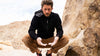 Man wearing black PAKA Breathe Zip Hoodie sitting on a rock in a sunlit desert landscape