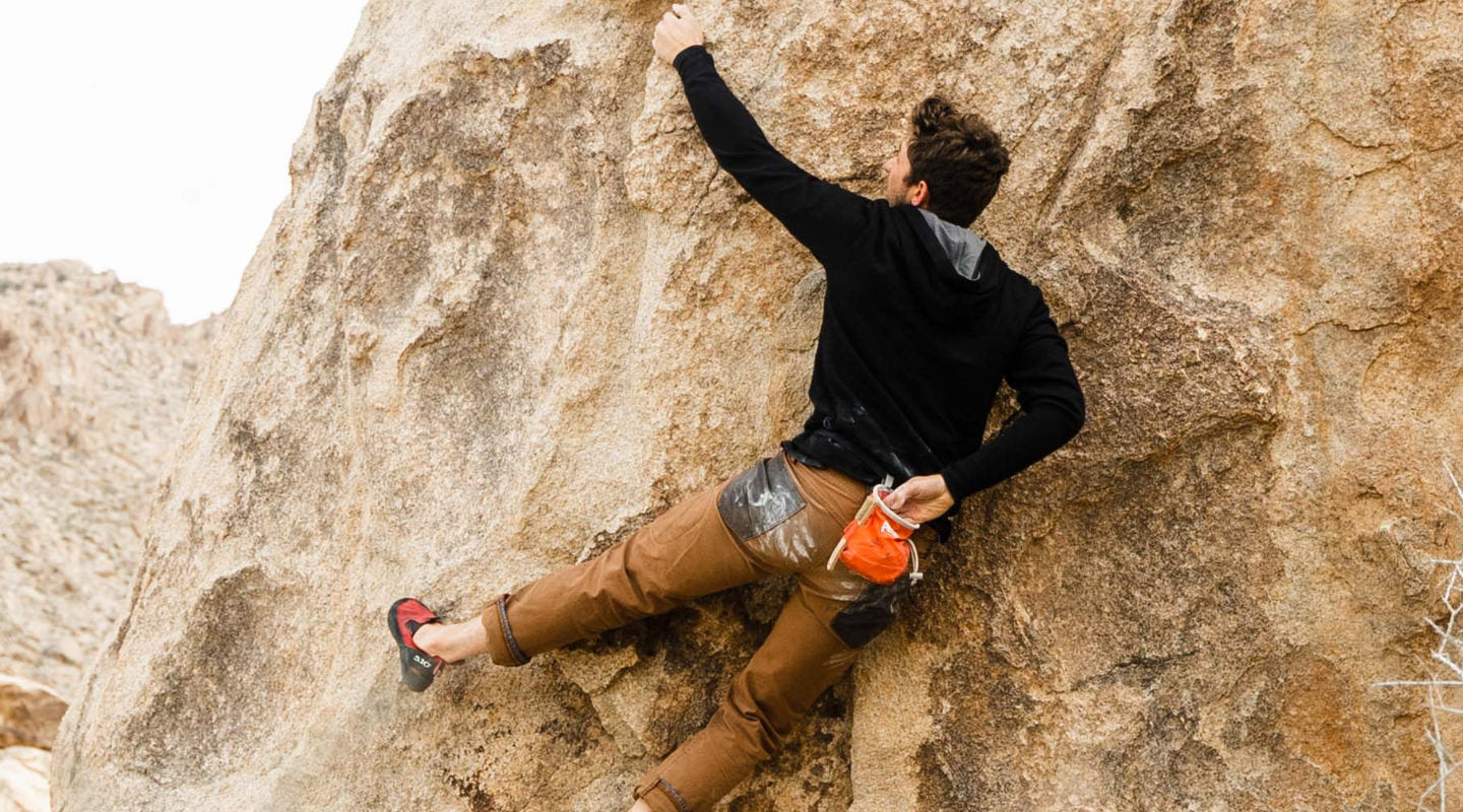 Man bouldering on a granite boulder wearing a black zip hoodie and brown pants with an orange chalk bag