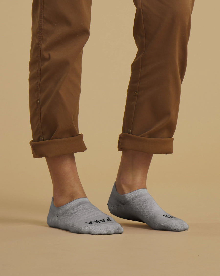 Angled front view of Essential No Show socks in light gray with low profile and PAKA logo on a beige background