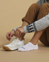 Seated person tying a beige sneaker while wearing white no-show PAKA socks on a neutral beige background