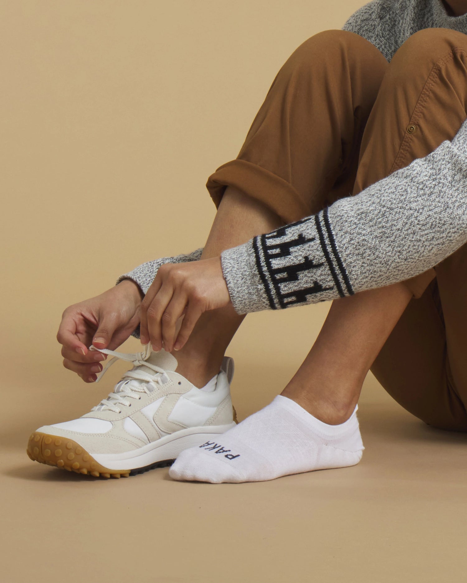 Seated person tying a beige sneaker while wearing white no-show PAKA socks on a neutral beige background