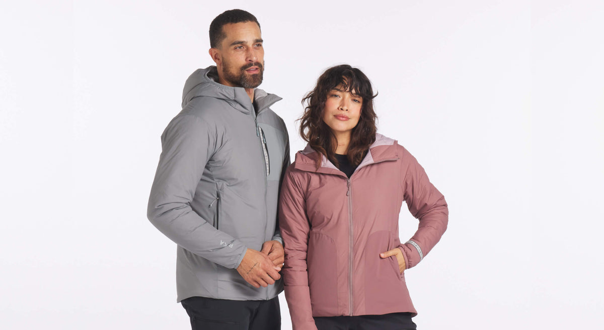 Three-quarter view of Apu PAKAFILL insulated jackets on models: man wearing gray hooded jacket and woman wearing dusty rose zip-front jacket with hand pocket on white background