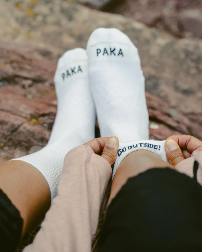 Front view of white Trail 1/4 crew socks on feet showing PAKA logo at cuff and go outside heel text, rocky outdoor background