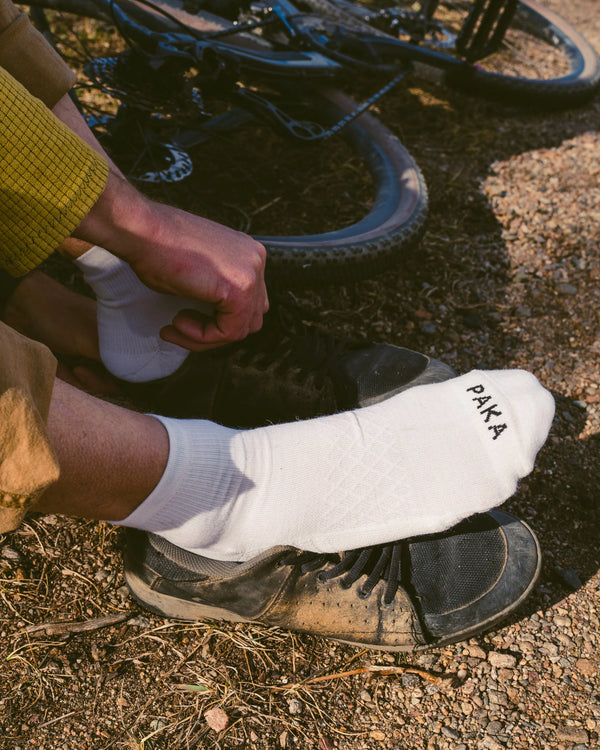 Side view of white Trail 1/4 crew socks on a person showing low crew height and PAKA logo on the toe, dirt trail with bike