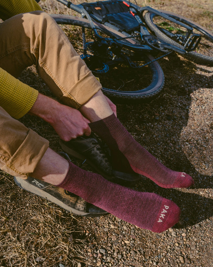 Model pulling on maroon 3/4 crew PAKA hiking socks showing PAKA logo on toes, sitting by a gravel trail with a mountain bike