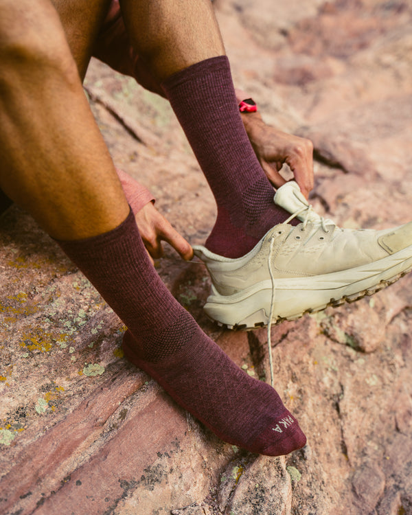 Angled view of Trail Crew Socks in burgundy showing crew height, textured knit and reinforced toe with PAKA logo on rocky background