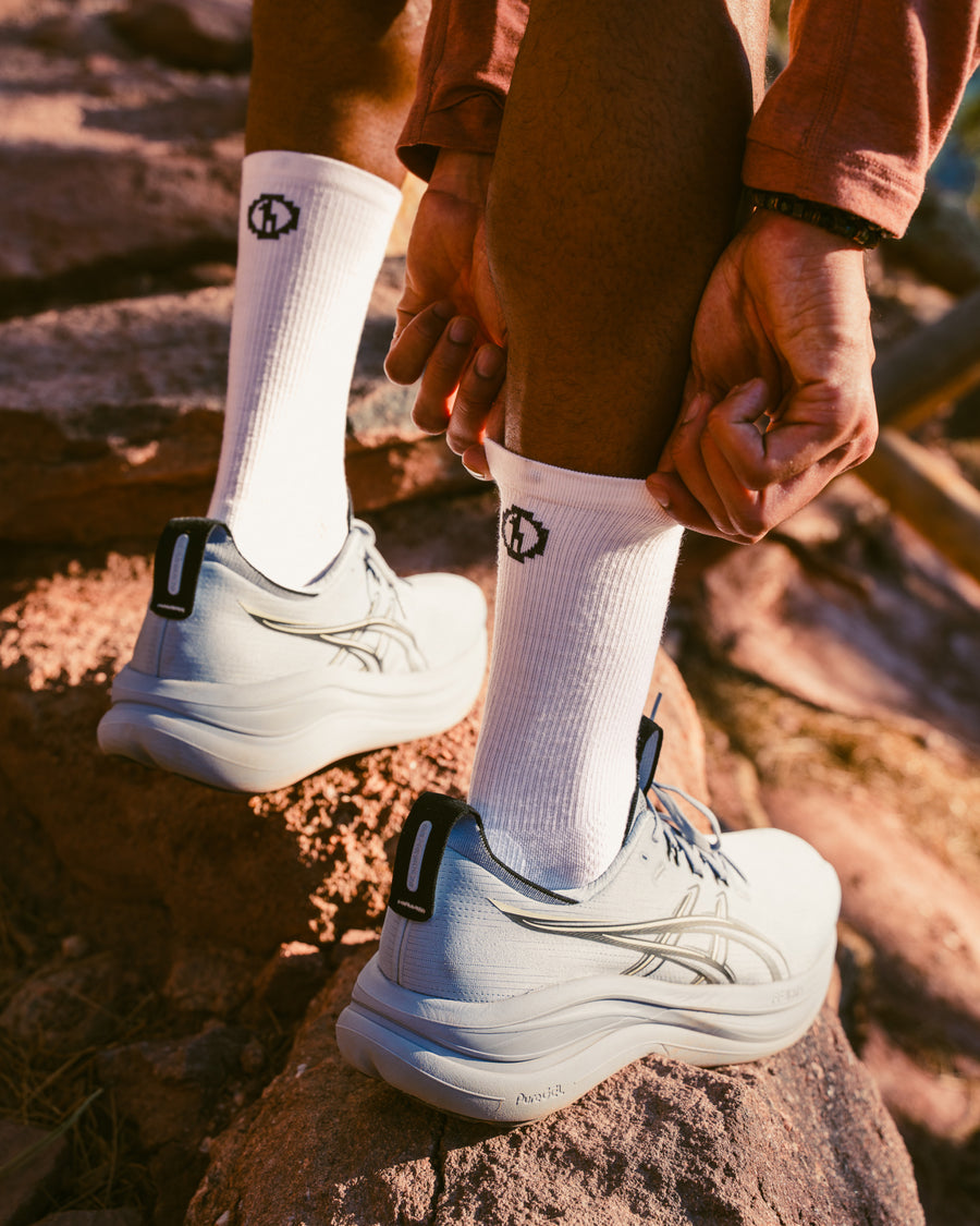 Rear three-quarter view of white Trail Crew socks showcasing mid-calf fit and ribbed texture on a model in hiking shoes on a rocky trail