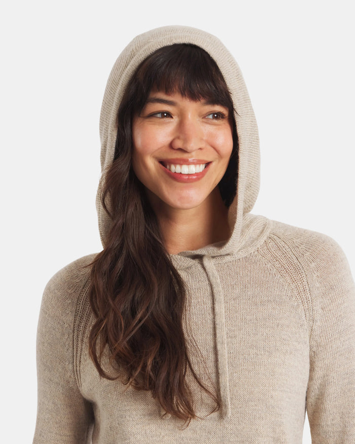 Front close-up of women’s alpaca hoodie in light beige on a smiling model, showing hood with drawstring and soft knit texture