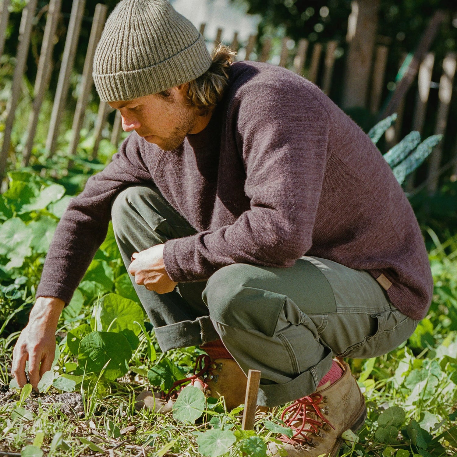 Man crouching in a garden wearing a brown alpaca crewneck sweater, olive pants, a knit beanie, and hiking boots