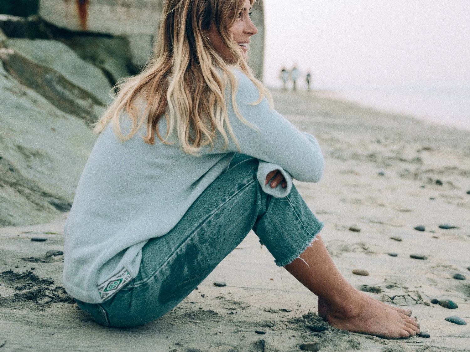 Woman sitting on a beach wearing a light blue alpaca crewneck, relaxed fit, smiling toward the ocean