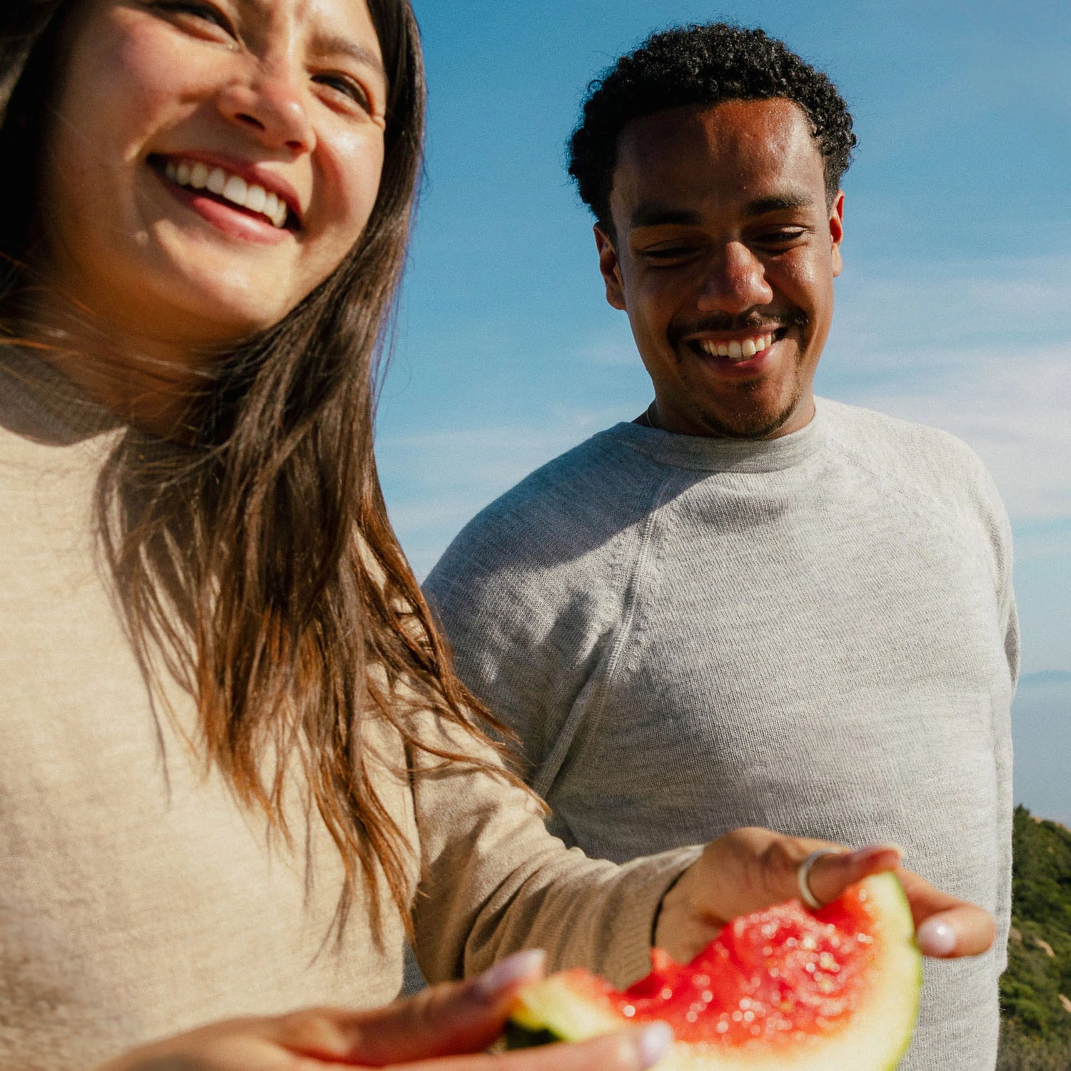 Two people smiling outdoors on a sunny day wearing alpaca crewneck sweaters against a blue sky