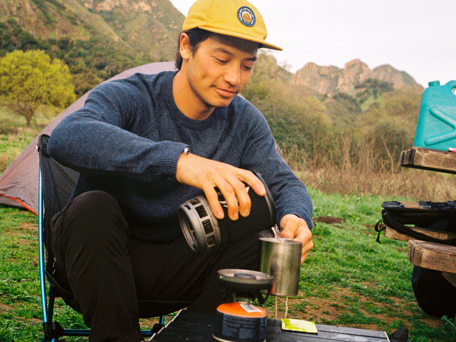 Man wearing navy men’s ultralight alpaca crewneck pouring water into a mug at a campsite with tent and hills behind him