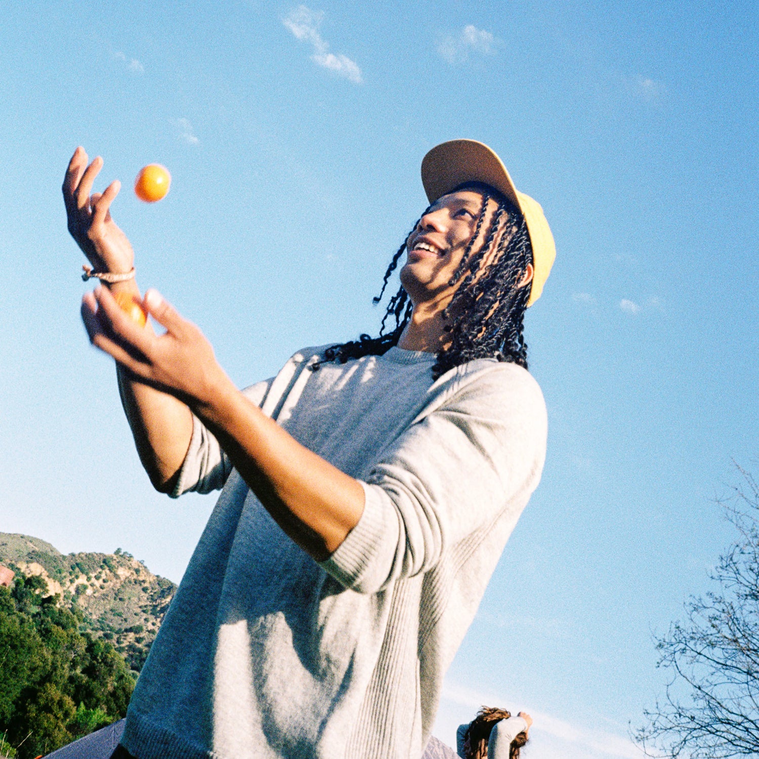 Young man tossing oranges while wearing a light gray Ultralight crewneck sweater outdoors against a blue sky