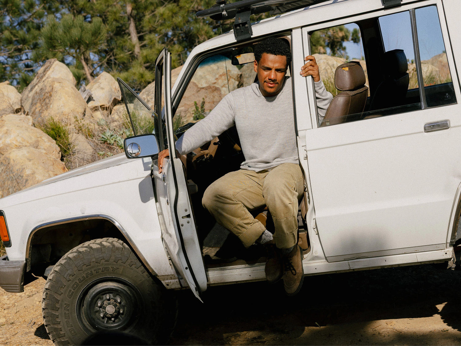 Man stepping out of a white truck wearing a light gray alpaca hoodie and khaki pants in a rocky outdoor setting