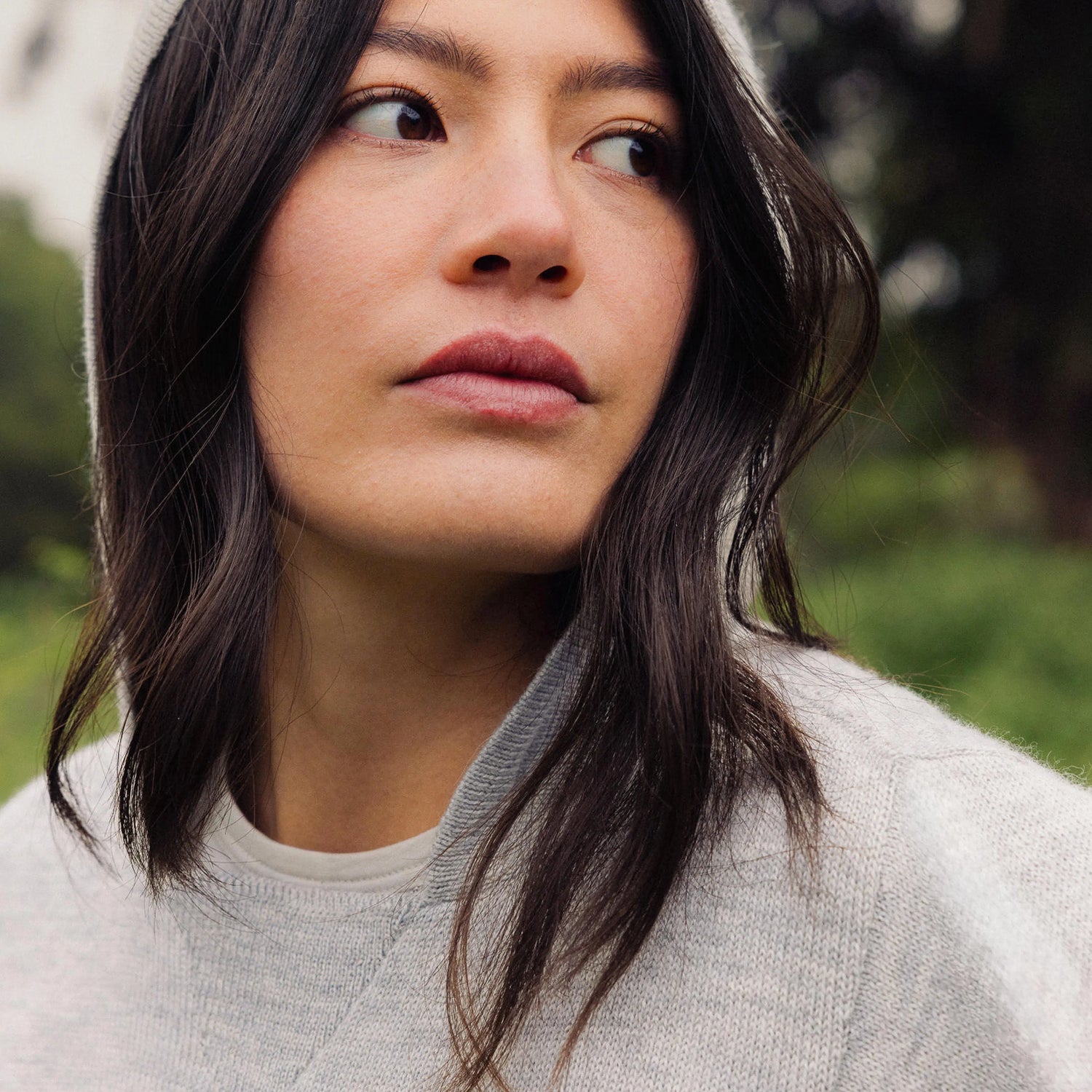 Close-up of a woman looking to the side wearing a light gray alpaca hoodie outdoors, showing the hood and knit texture