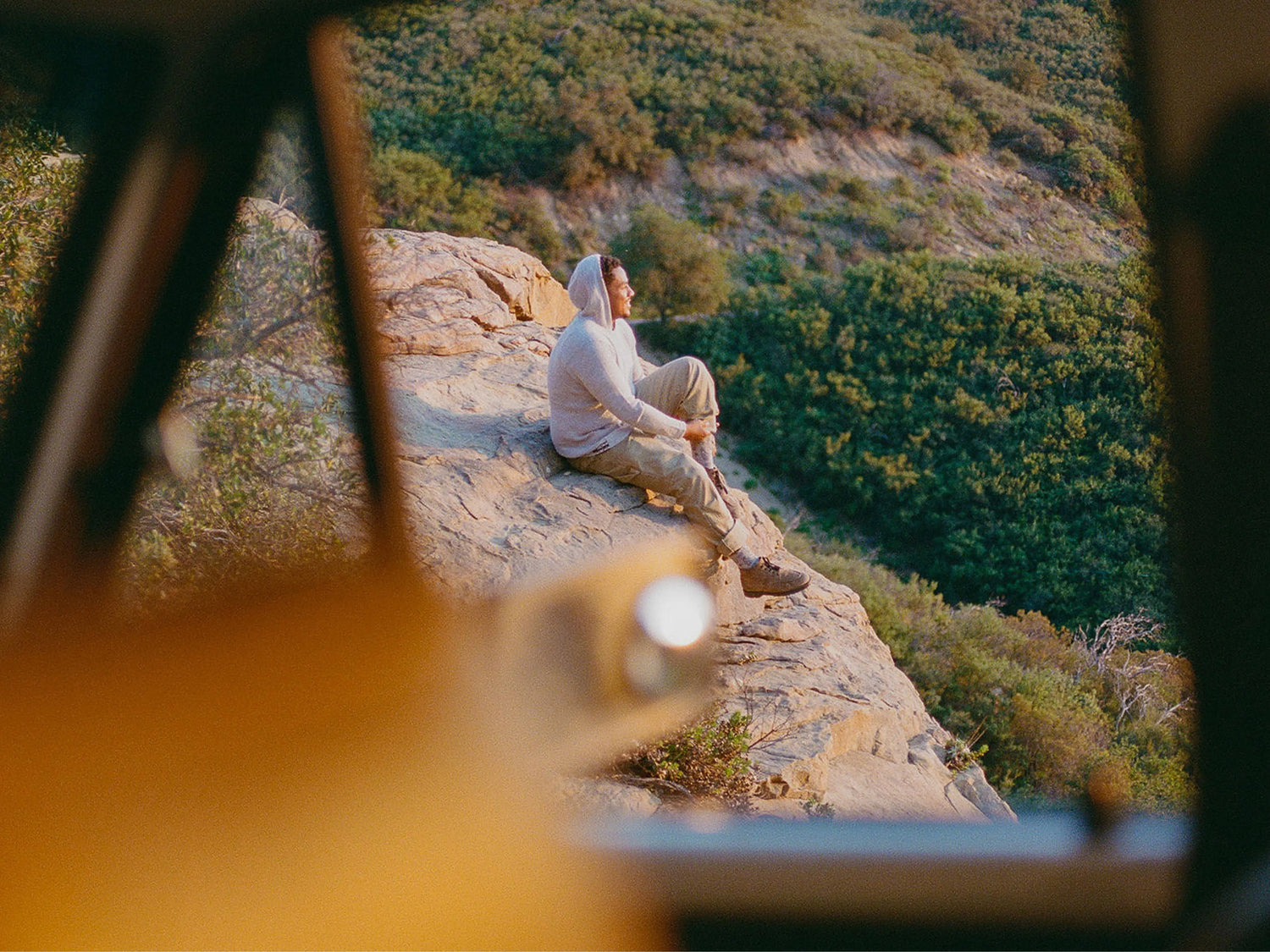 Person wearing an off-white alpaca hoodie sitting on a rocky cliff, looking over a green canyon at golden-hour light