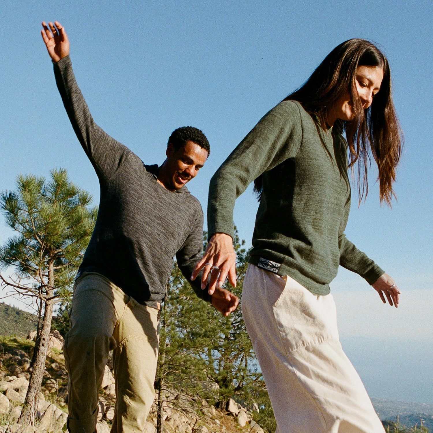 Two people wearing alpaca hoodies holding hands and balancing on rocks with pine trees and a clear blue sky background