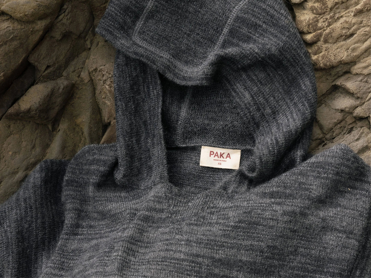 Close-up of charcoal alpaca hoodie showing soft ribbed knit, hood and PAKA label on a rocky background