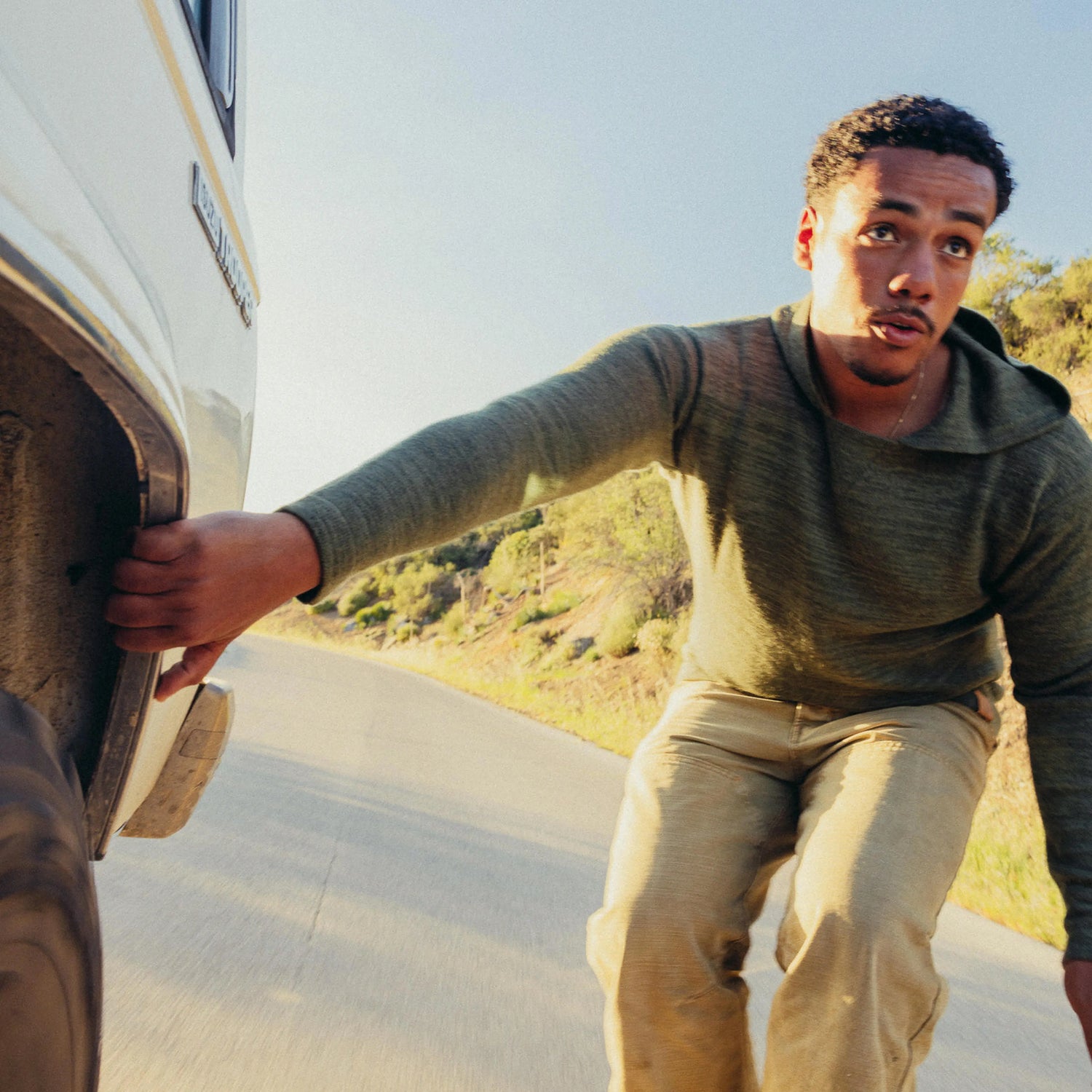 Man leaning on a car wearing an olive green unisex alpaca hoodie, relaxed fit, sunlit roadside hills in background