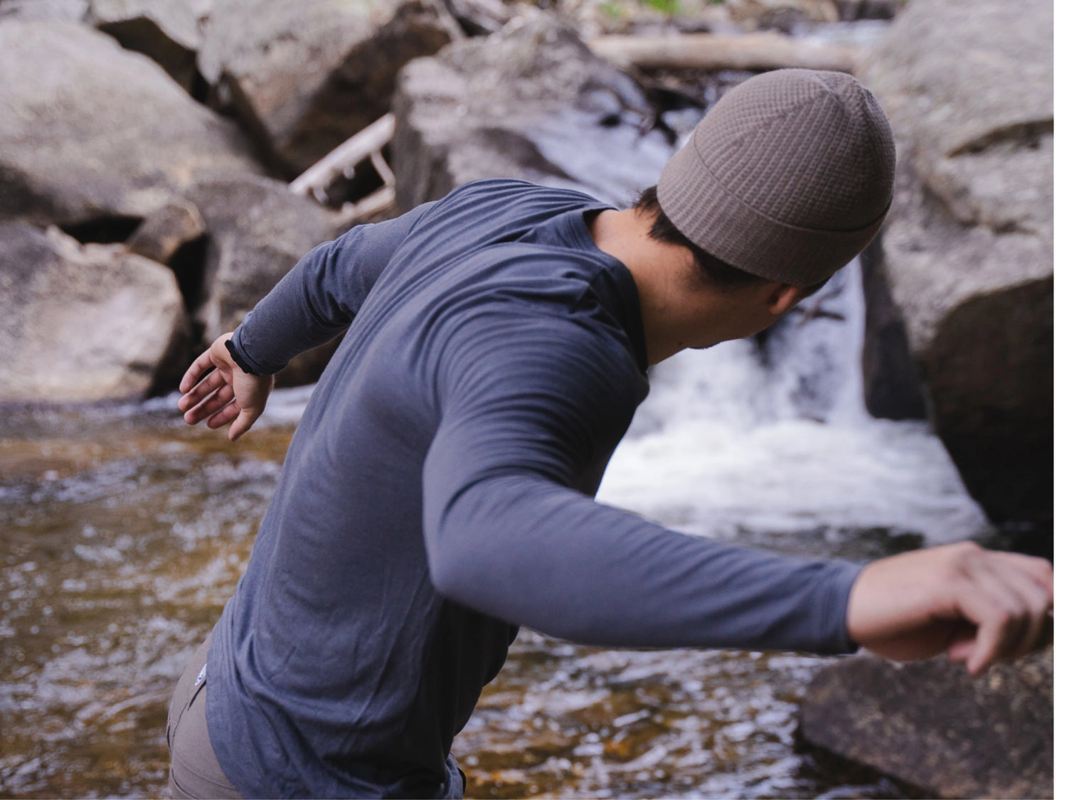 Man balancing on rocks by a stream wearing a charcoal Mens Aire long sleeve alpaca shirt