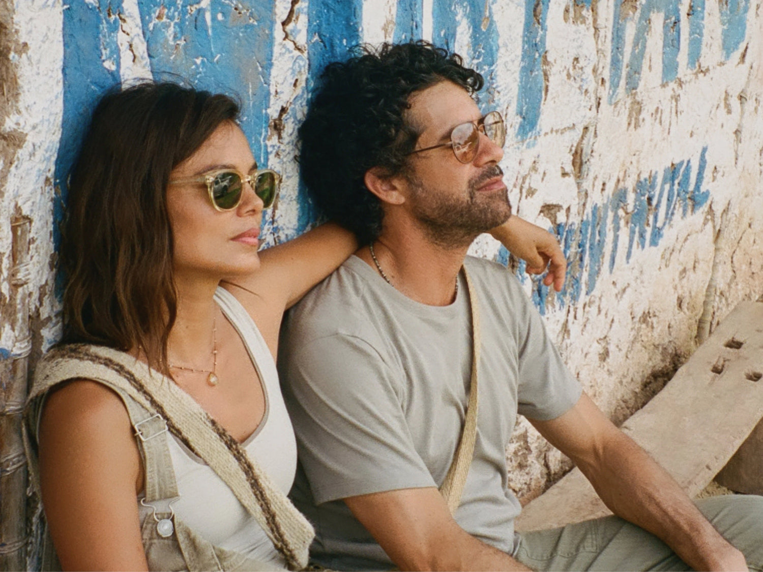 Man in a light gray slim-fit tee and sunglasses leaning against a weathered blue-and-white wall beside a woman, relaxed outdoor pose