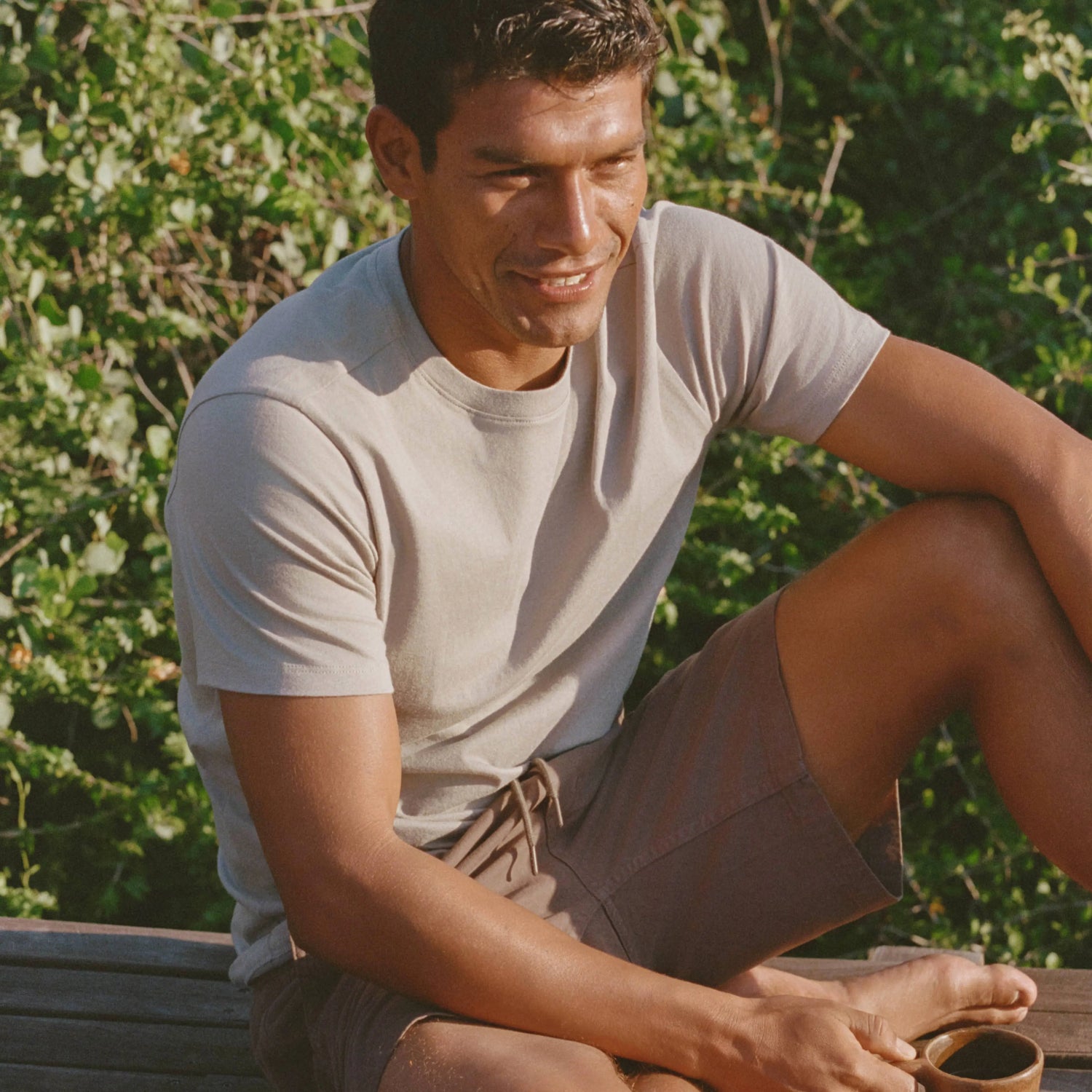 Man sitting outdoors smiling, wearing a light beige Men’s Essential Tee in a relaxed fit against a leafy background