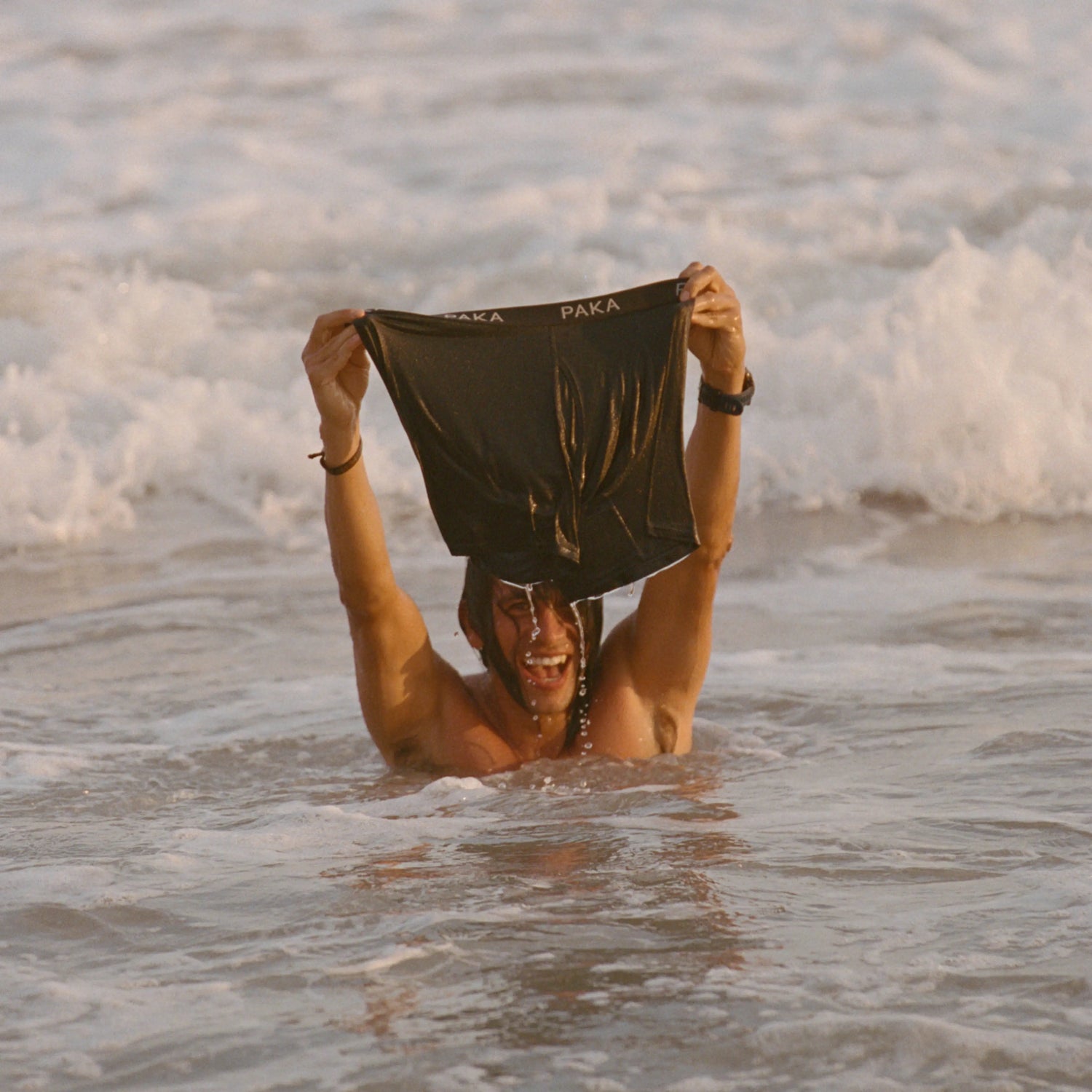 Man in the ocean holding up black PAKA boxer briefs by the waistband against foamy waves
