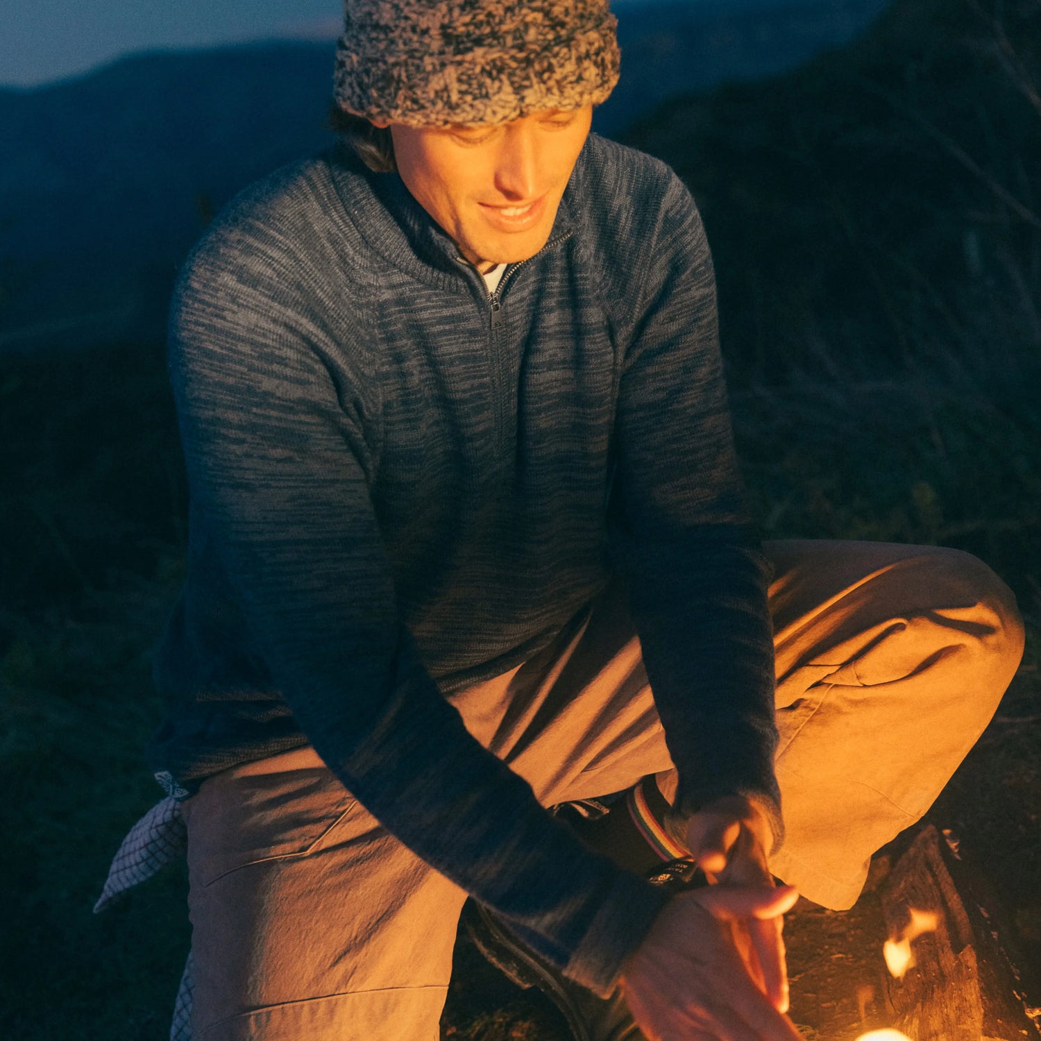 Man wearing navy alpaca quarter-zip sweater warming his hands by a campfire at dusk