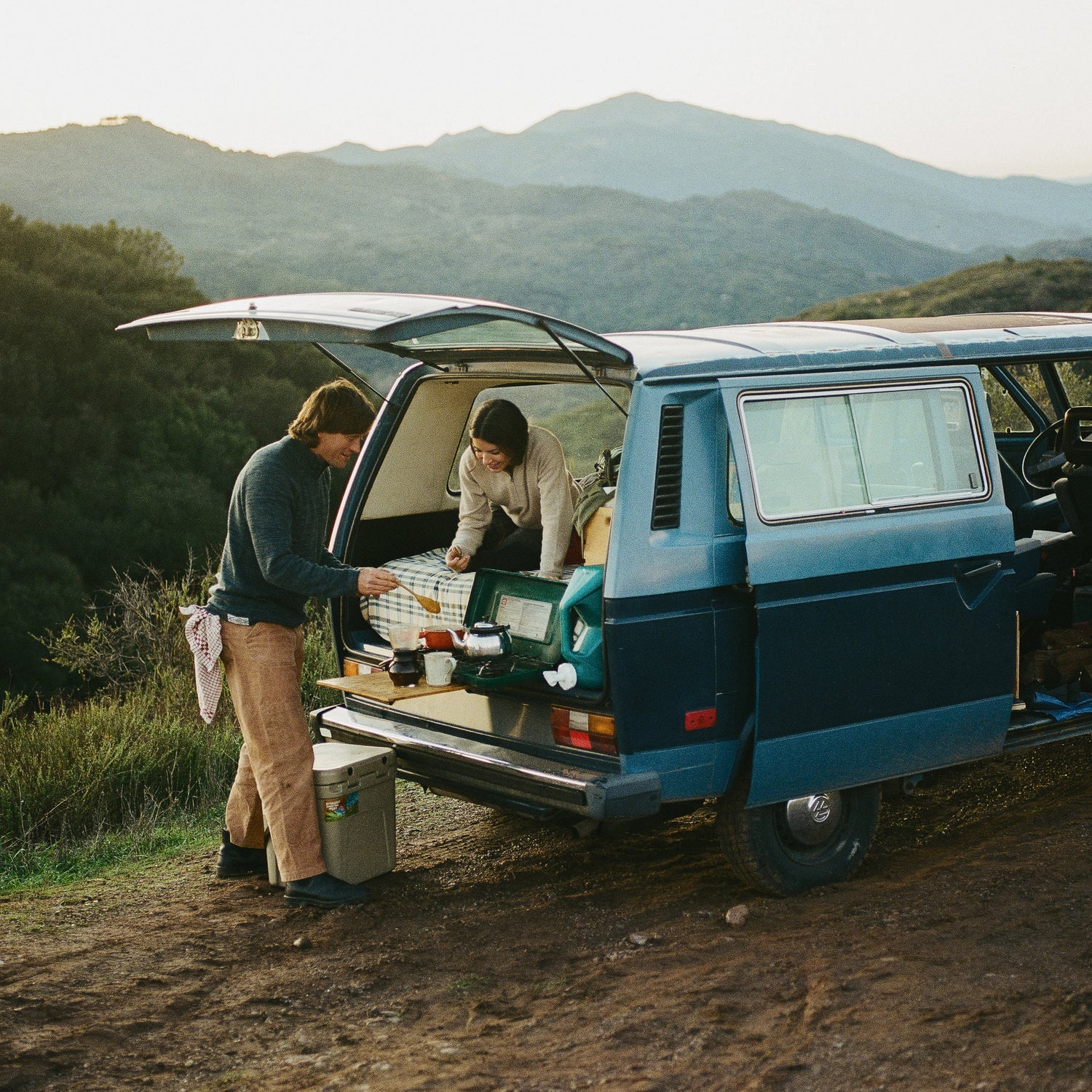 Two people loading a blue camper van, one wearing a light alpaca quarter-zip sweater, mountains in the background