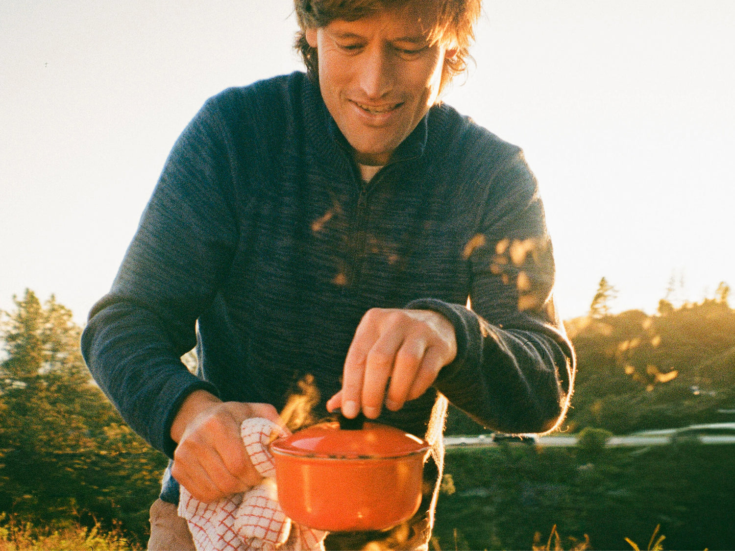 Person cooking over a campfire at sunset wearing a navy alpaca wool quarter-zip sweater (relaxed fit) by a lake
