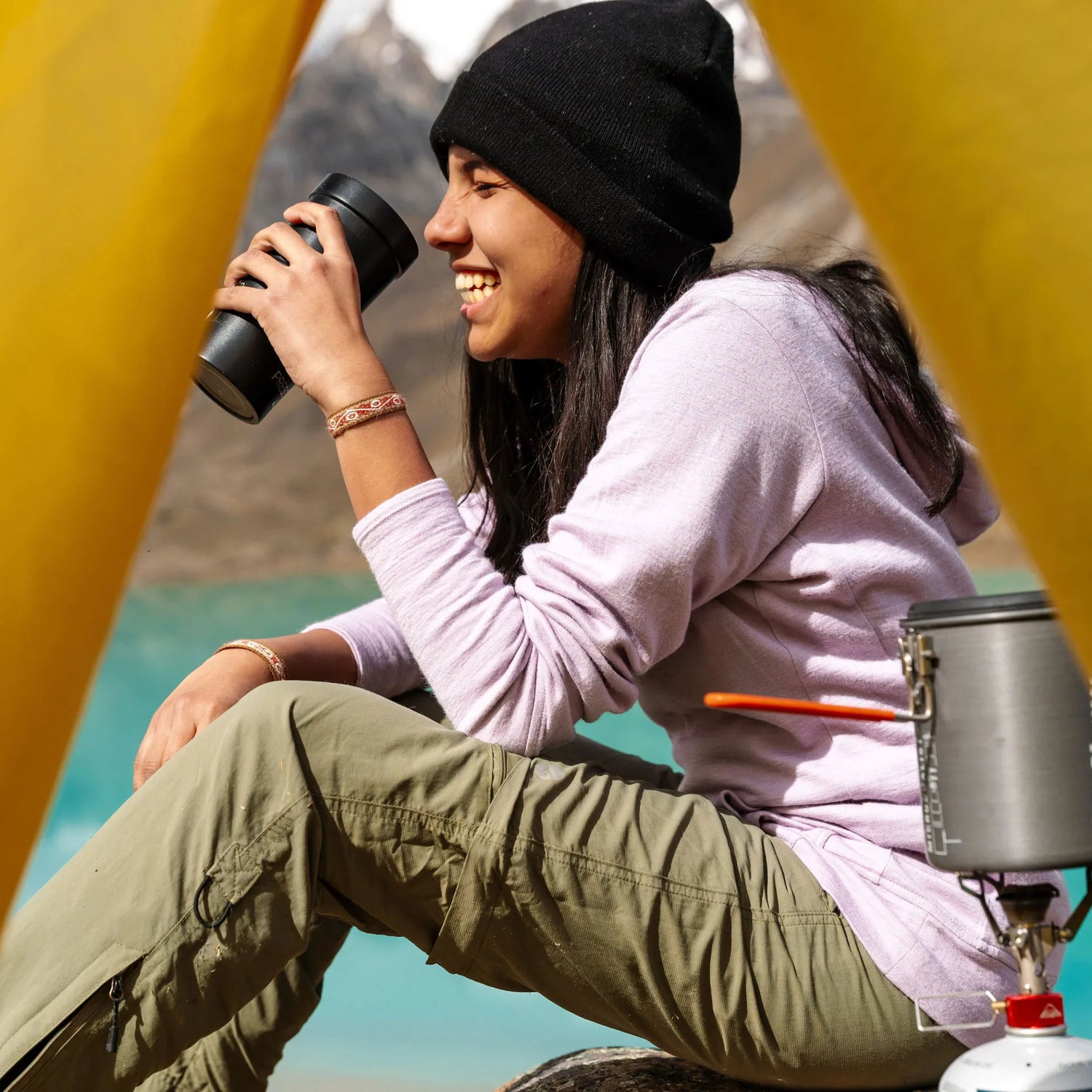 A smiling woman holding a cup of coffee and wearing an alpaca bracelet in front of a beautiful lake
