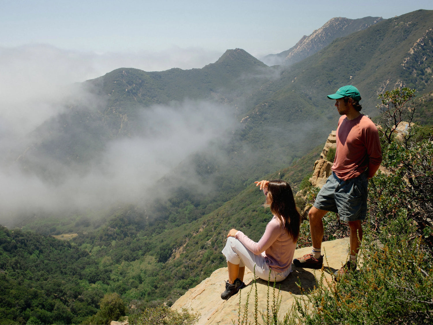 Man wearing rose Mens Sol Crew long-sleeve UPF shirt relaxed fit, standing on cliff overlooking foggy mountains